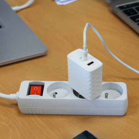 Chargeur secteur blanc branché sur une multiprise et relié à un câble USB-C, dans un environnement de bureau. Aucun marquage vis