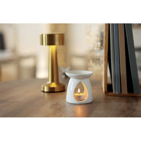 Brûleur de parfum en céramique blanche posé sur un meuble en bois, avec une bougie chauffe-plat allumée visible dans son ouvertu