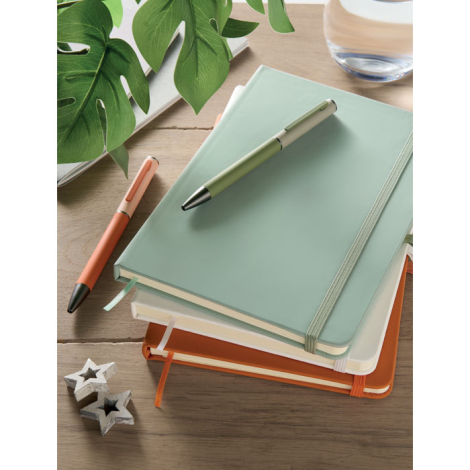 Mise en scène de plusieurs cahiers A5 empilés dans différents coloris, accompagnés de stylos, avec un cahier vert clair au premi