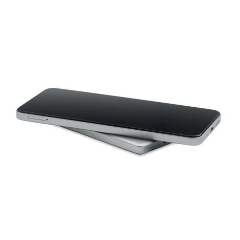 Smartphone noir posé sur la batterie externe argentée, légèrement décalée sous l’appareil.