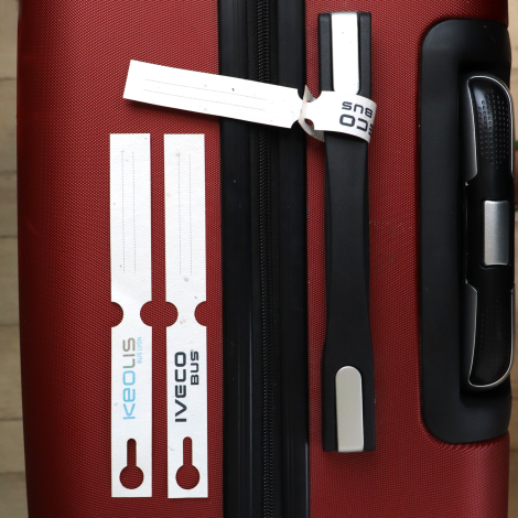 Deux étiquettes de bagage blanches personnalisées avec les logos IVECO BUS et KEOLIS, présentées à plat sur une valise rouge. Un