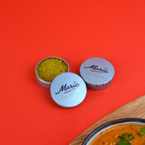 Trois petites boîtes métalliques rondes personnalisées Marie, dont une ouverte contenant du curry jaune, disposées sur fond roug