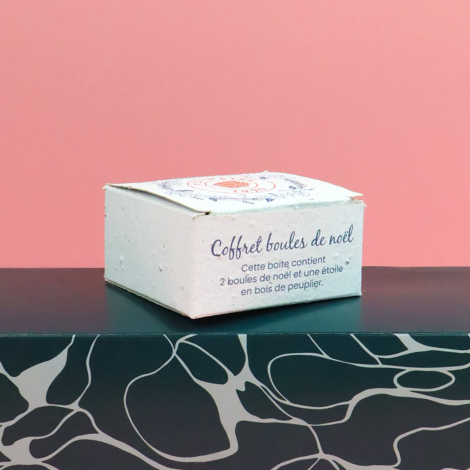 Boîte en papier ensemencé personnalisée « Coffret boules de Noël », présentée sur fond rose avec un support noir à motifs blancs