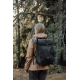 Femme vue de trois quarts dos dans un environnement boisé, portant un sac à dos noir à rabat fermé par une boucle centrale, avec