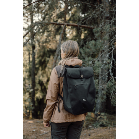 Femme vue de trois quarts dos dans un environnement boisé, portant un sac à dos noir à rabat fermé par une boucle centrale, avec