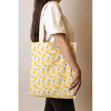 Cabas Nina écru à motifs citrons jaunes et feuilles vertes, porté à l'épaule par une femme, avec longues anses écrues et étiquet