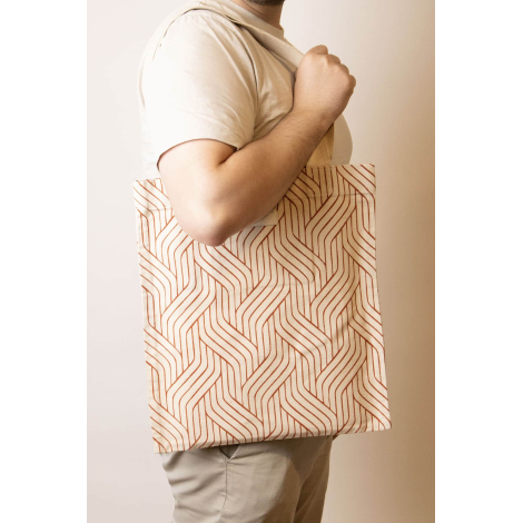 Cabas Nina écru à motifs géométriques orange, porté à l'épaule par un homme, avec longues anses écrues et étiquette frontale.