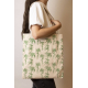 Cabas Nina écru à motifs palmiers verts, porté à l'épaule par une femme, avec longues anses écrues et étiquette rectangulaire su