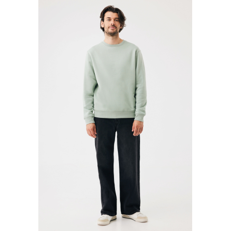 Homme debout de face portant un sweat vert sauge clair à col rond et manches longues, associé à un jean noir ample et des basket