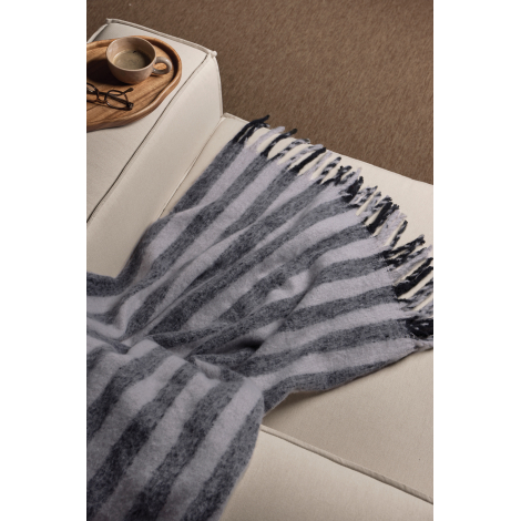 Couverture rayée gris foncé et gris clair étendue sur un canapé beige, avec ses longues franges apparentes. Un plateau en bois p