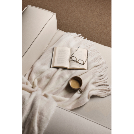 Couverture beige clair et écru drapée sur un canapé crème, avec ses franges épaisses sur le côté. Un livre ouvert, des lunettes 