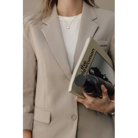 Femme vêtue d'un blazer beige et d'un haut blanc, tenant contre elle une bouteille isotherme noire mate avec manchon en silicone