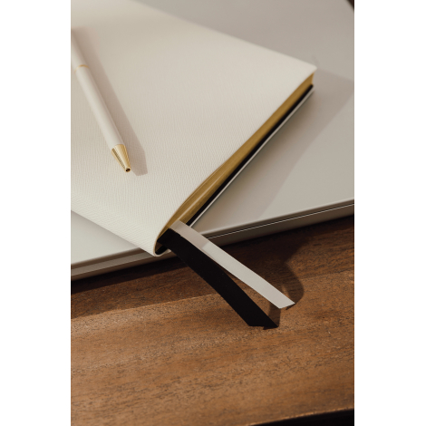 Gros plan sur un carnet beige à couverture texturée et tranches dorées, accompagné d'un stylo blanc à pointe dorée. Deux rubans 