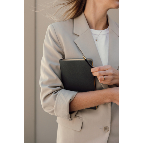 Femme vêtue d'un blazer beige tenant contre elle un carnet noir à couverture texturée, tout en maintenant un stylo bille noir à 