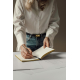 Femme en chemise blanche et jean bleu écrivant avec un stylo clair dans un carnet ouvert aux pages lignées et tranches dorées, p