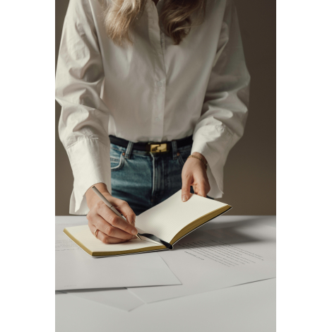 Femme en chemise blanche et jean bleu écrivant avec un stylo clair dans un carnet ouvert aux pages lignées et tranches dorées, p