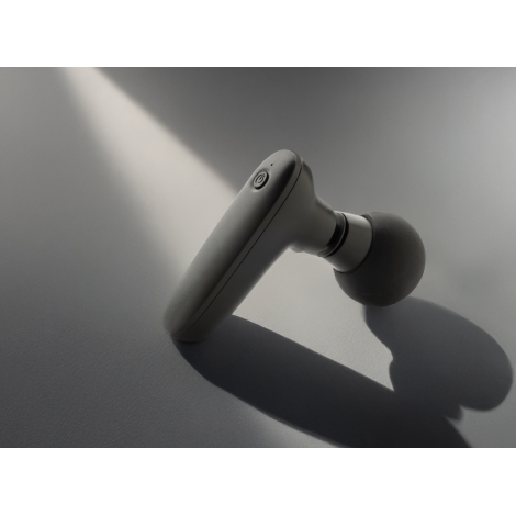 Pistolet de massage noir posé en biais sur surface grise avec jeu d’ombre, neutre sans marquage