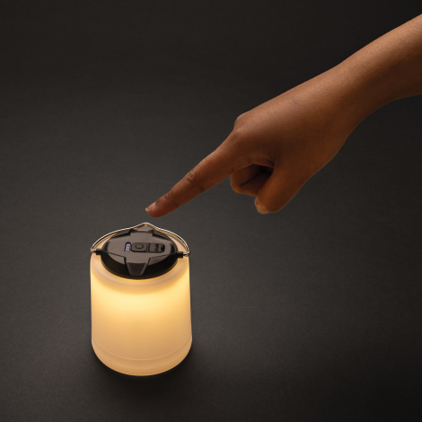 Lampe de table allumée en lumière chaude, posée sur une surface sombre avec une main sur le bouton supérieur.