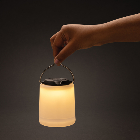 Lampe de table allumée en lumière chaude, tenue par son anse métallique.