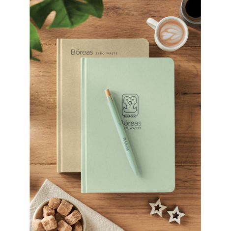 Deux carnets beige et vert avec stylo et tasse ; marquage visible sur la couverture du carnet vert et sur le stylo.