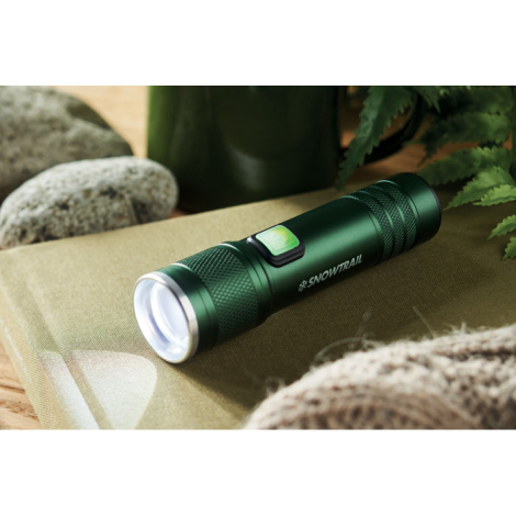 Lampe torche en aluminium vert foncé posée sur textile beige avec pierres et feuillage ; marquage visible sur la face latérale.