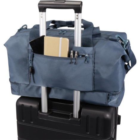Sac de voyage bleu fixé sur la poignée télescopique d’une valise, poche avant contenant un carnet et des stylos.
