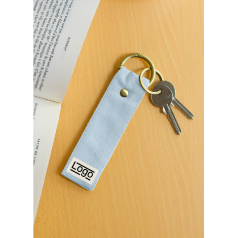 Porte-clés bleu clair posé sur bois clair, près d’un livre ouvert et d’un trousseau, marquage avant.