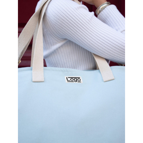 Sac bleu clair porté à l’épaule par une personne en blanc, marquage centré.