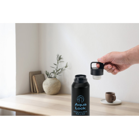 Bouteille noire avec bouchon dévissé tenue par une main, posée sur une table claire ; marquage visible sur la face avant.