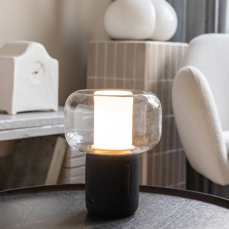 Lampe haut-parleur allumée posée sur une table en intérieur, diffuseur en verre et socle noir avec boutons latéraux visibles, sa