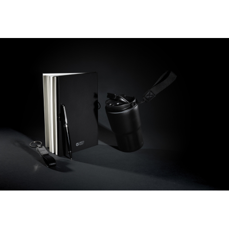 Mise en scène sur fond sombre avec carnet légèrement ouvert, stylo à proximité, porte-clés au premier plan et mug en arrière-pla