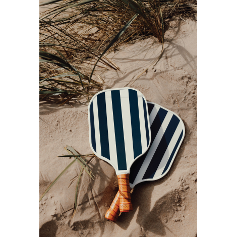 Deux raquettes de pickleball en bois à motif rayé bleu et blanc posées sur le sable avec une balle, présentées en extérieur et s