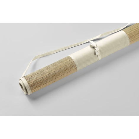 Natte de plage en paille naturelle présentée enroulée sur fond clair, vue de biais avec bande textile d’enroulage et sangle de t