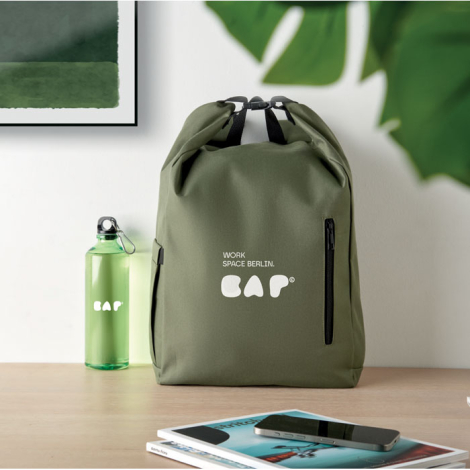 Sac à dos vert posé sur un bureau, vue de face avec marquage visible centré sur la face avant au-dessus de la poche zippée.