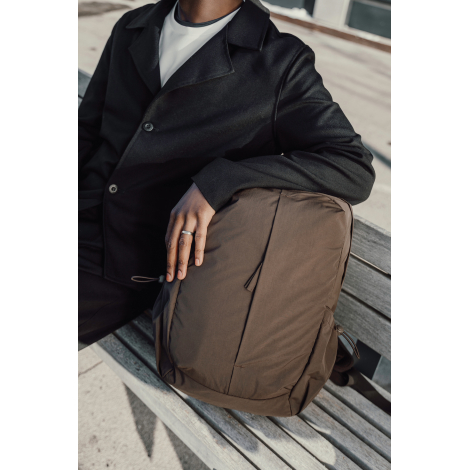 Sac à dos marron tenu par une personne assise sur un banc, en situation d’usage, sans personnalisation apparente.