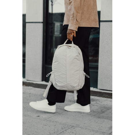 Sac à dos beige porté à la main par une personne en extérieur, illustrant le transport, sans personnalisation visible.