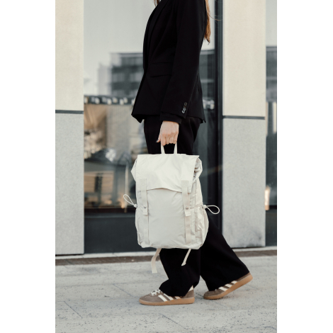 Sac à dos beige porté à la main par une personne en extérieur, illustrant le transport, sans personnalisation visible.