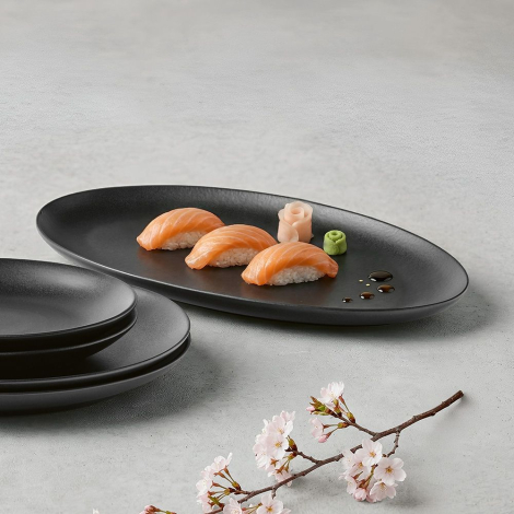 Plat ovale en céramique noire utilisé pour présenter des aliments avec assiettes empilées à proximité, sans marquage apparent