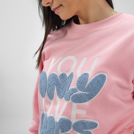 Gros plan sur une femme portant un sweat femme à col rond rose avec un grand marquage graphique texturé visible sur la poitrine.