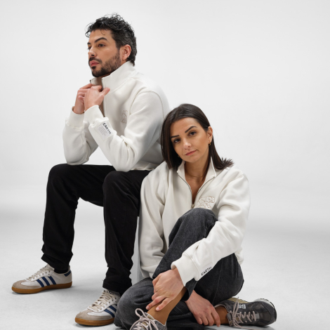 Un homme et une femme portent chacun un sweat unisexe blanc à col montant zippé dans un studio photo. Une broderie est visible s